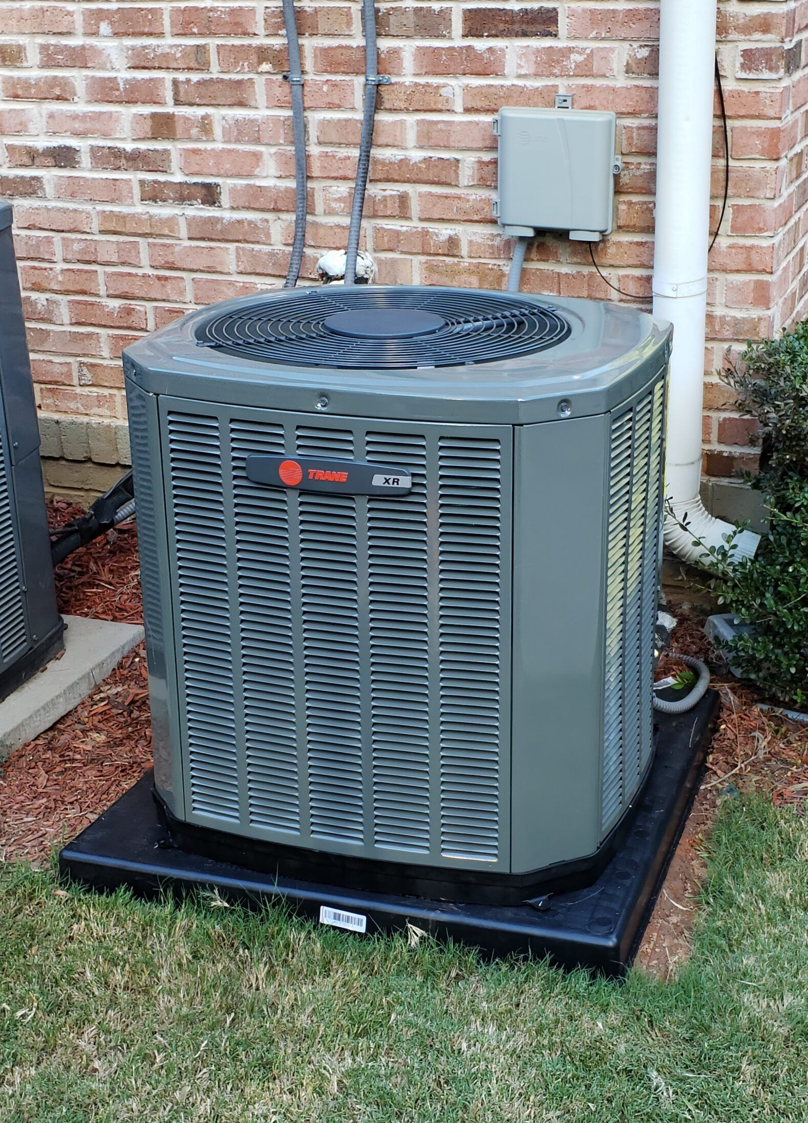 Outdoor HVAC condenser unit beside a brick house wall on a black pad, with grass and utility box nearby