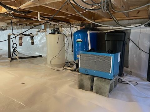 Under-house crawl space with blue HVAC unit, water heater, and exposed pipes and wiring on plastic sheeting.