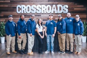 Eight people in blue jackets and T-shirts standing in front of a wooden wall with the word “CROSSROADS.”