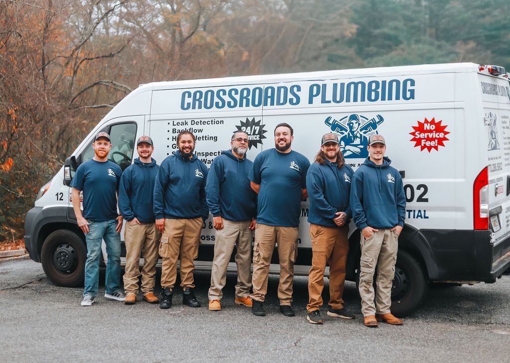 Plumbing crew posing in front of a white Crossroads Plumbing van on a gravel lot.
