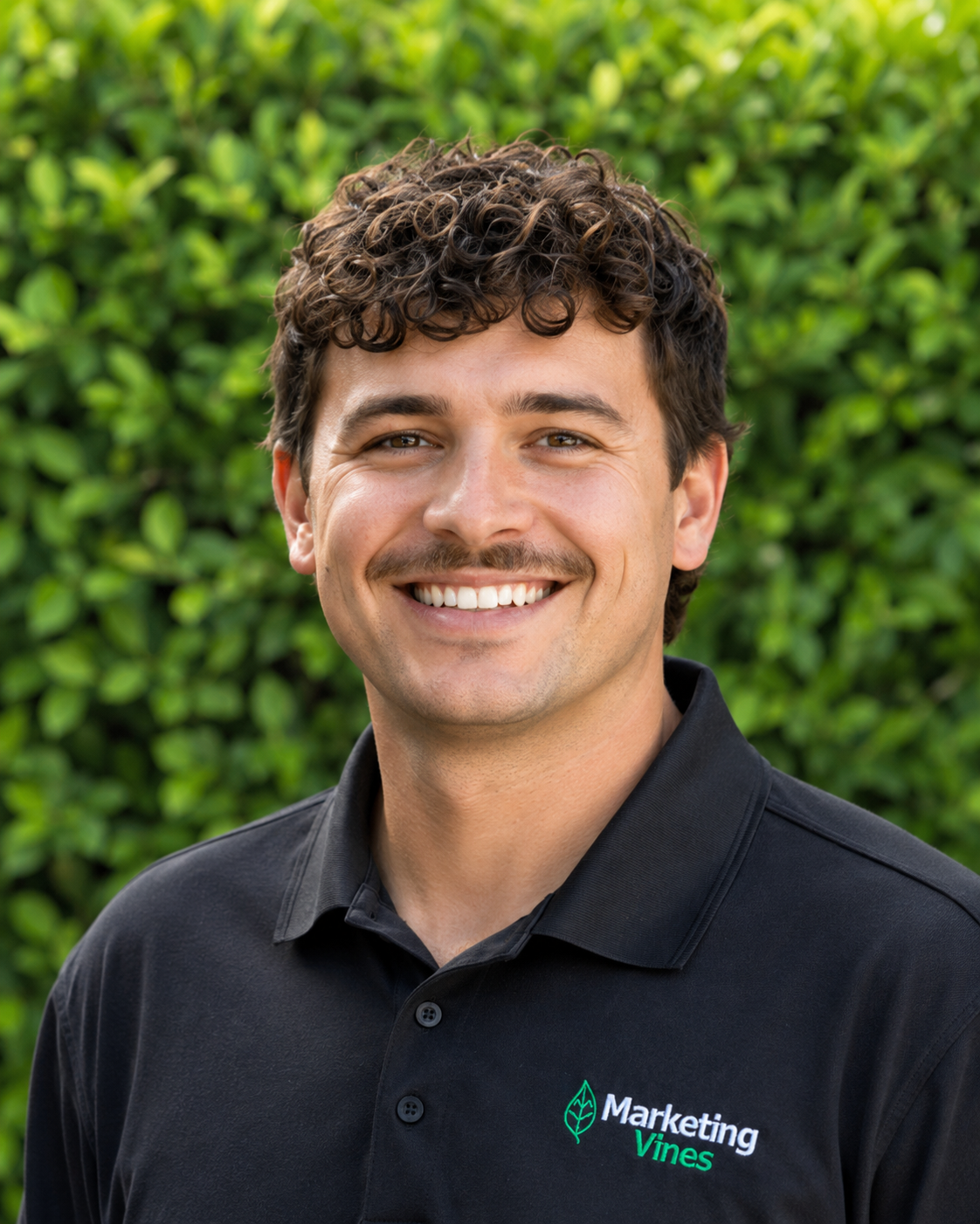 Mason Whitaker, Director of Client Strategy & Operations for Home Service Businesses Smiling man in a black polo shirt standing outdoors in front of green foliage