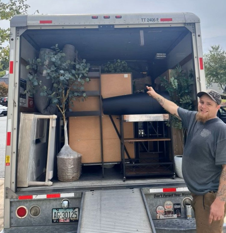 Mover pointing at packed truck with boxes, furniture, and a potted tree inside
