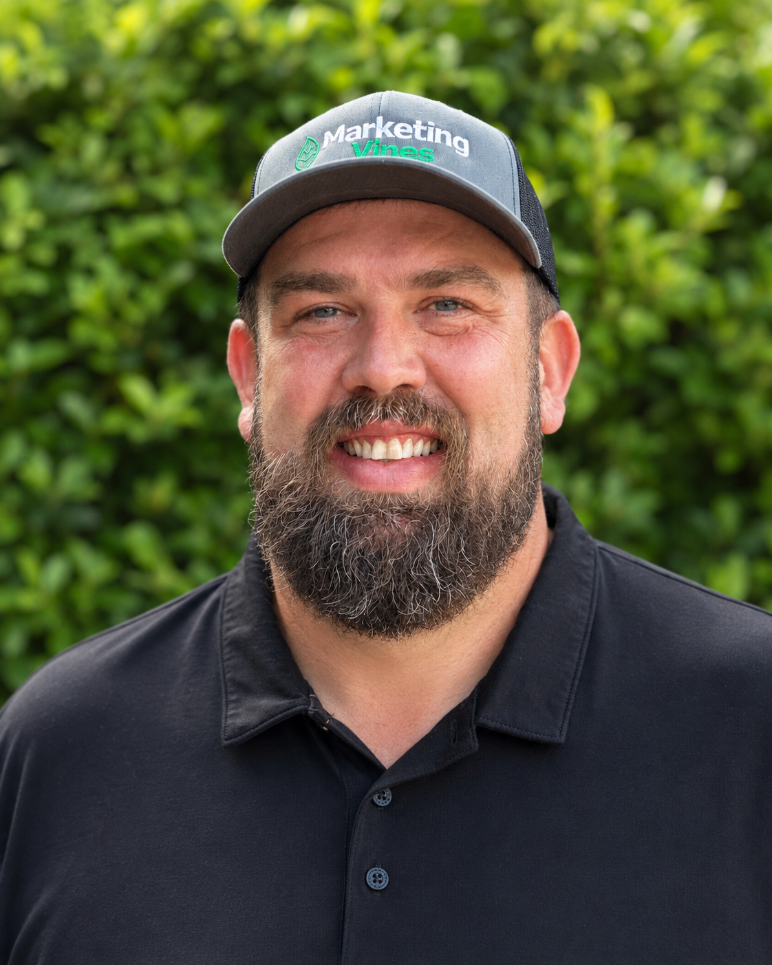 Timothy Bennett, Marketing Vines Founder Man in a black polo and cap smiling outdoors against a green leafy background