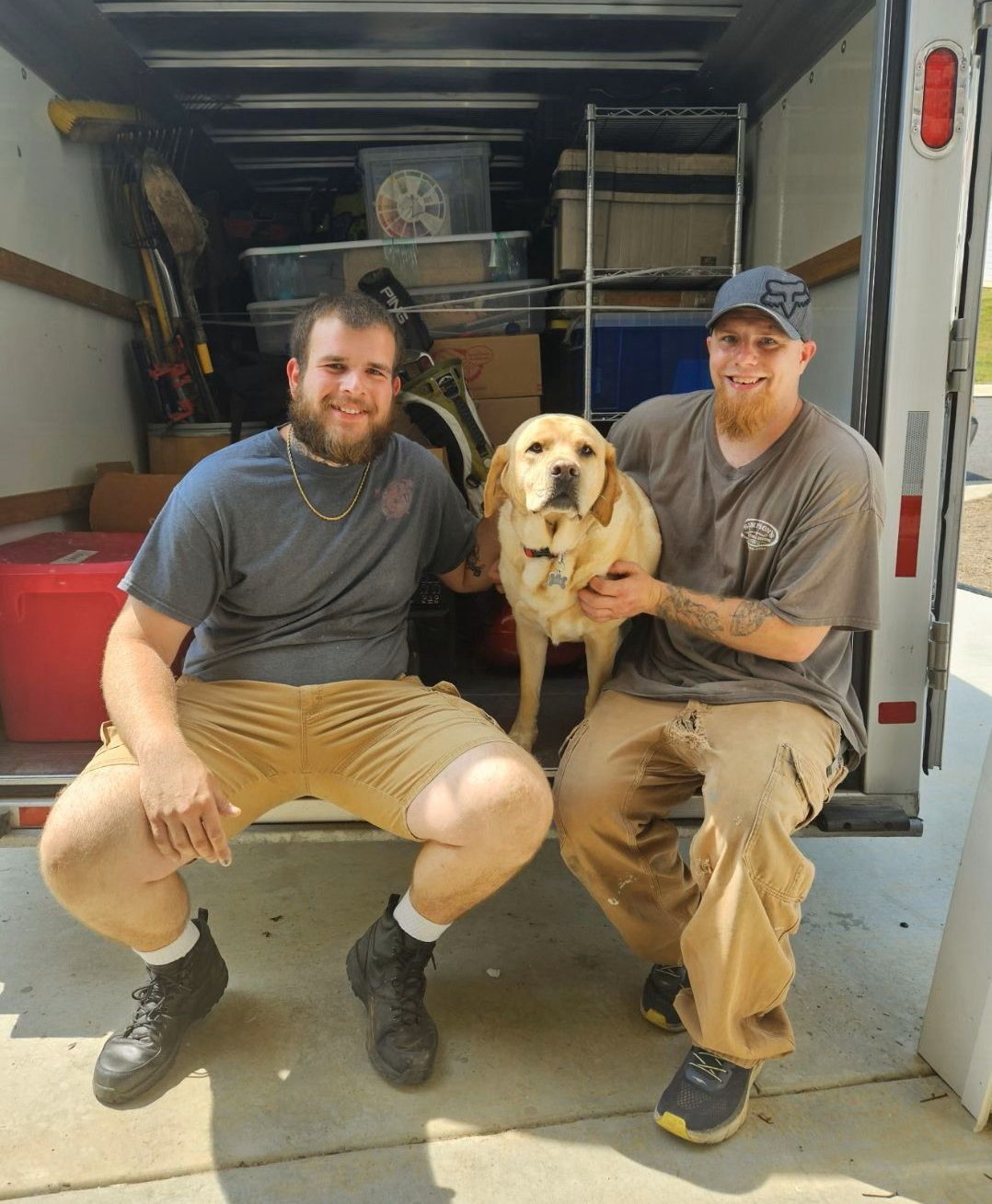 Two men kneel beside a yellow dog in the back of a moving truck, smiling at the camera.