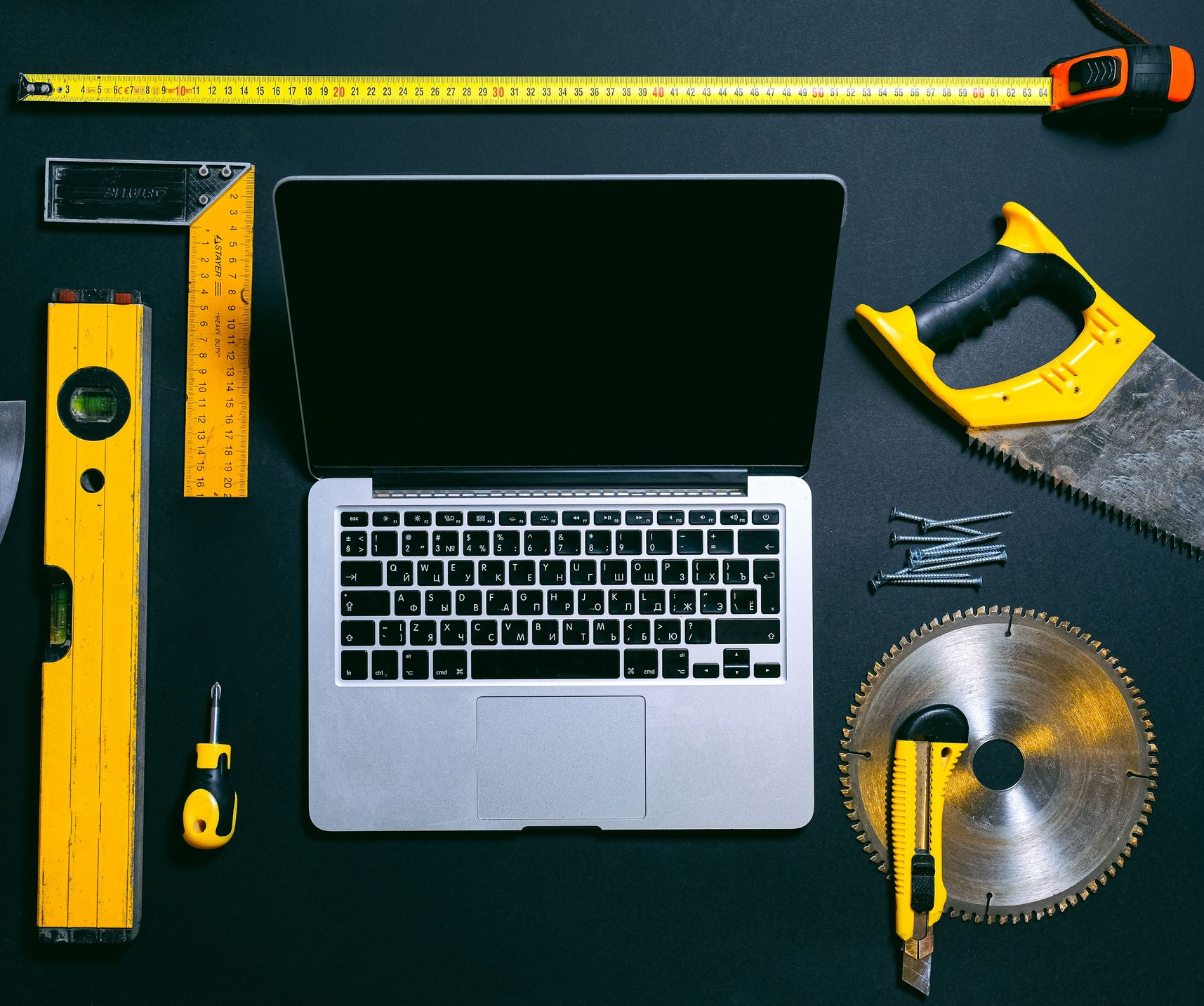 Laptop with yellow measuring tools, tape measure, and saw arranged on a dark work surface