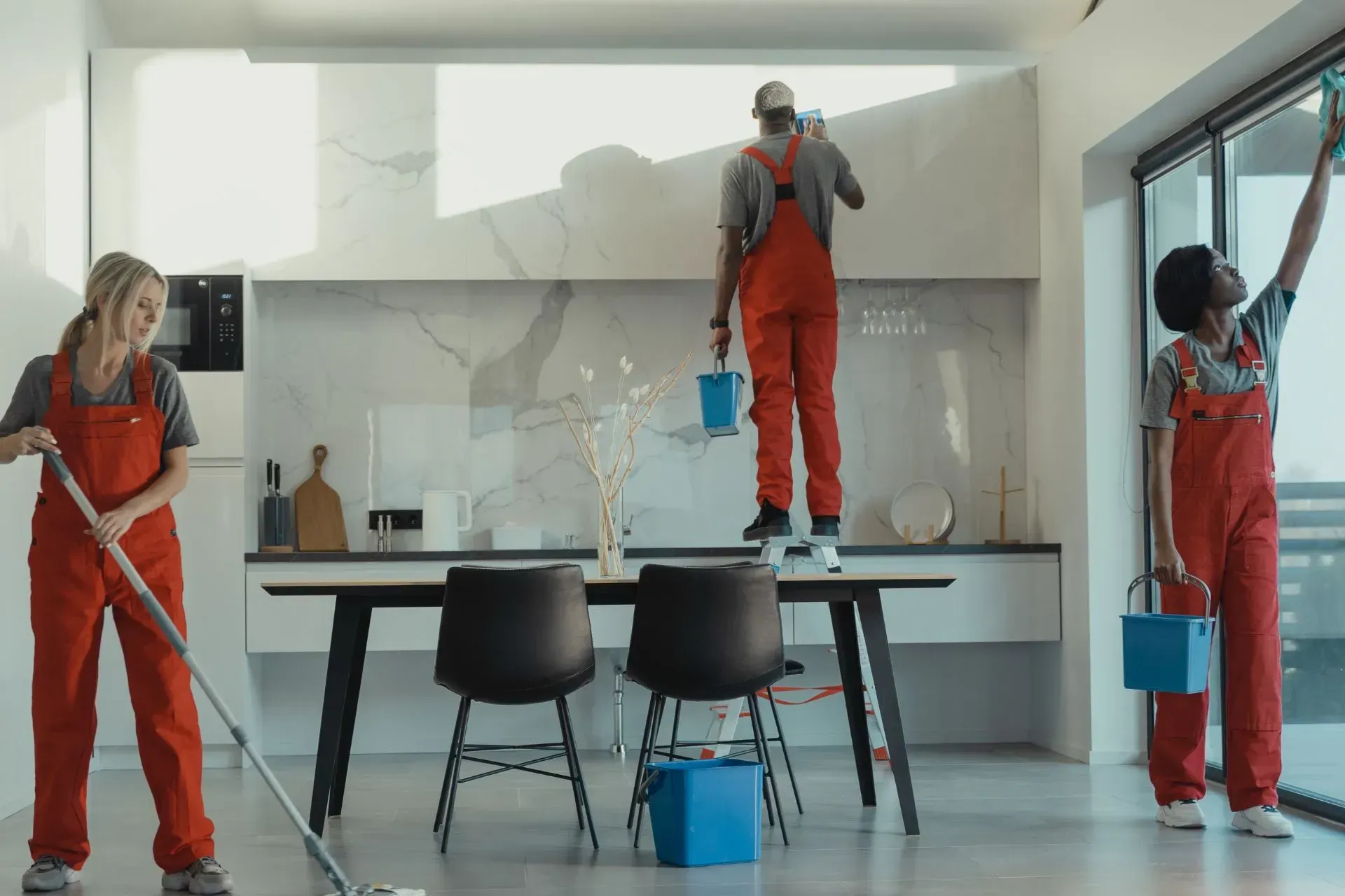 Office Cleaning Services Henderson NV-Three people in red jumpsuits cleaning a bright kitchen. One mops, one paints high up, and another washes a window.