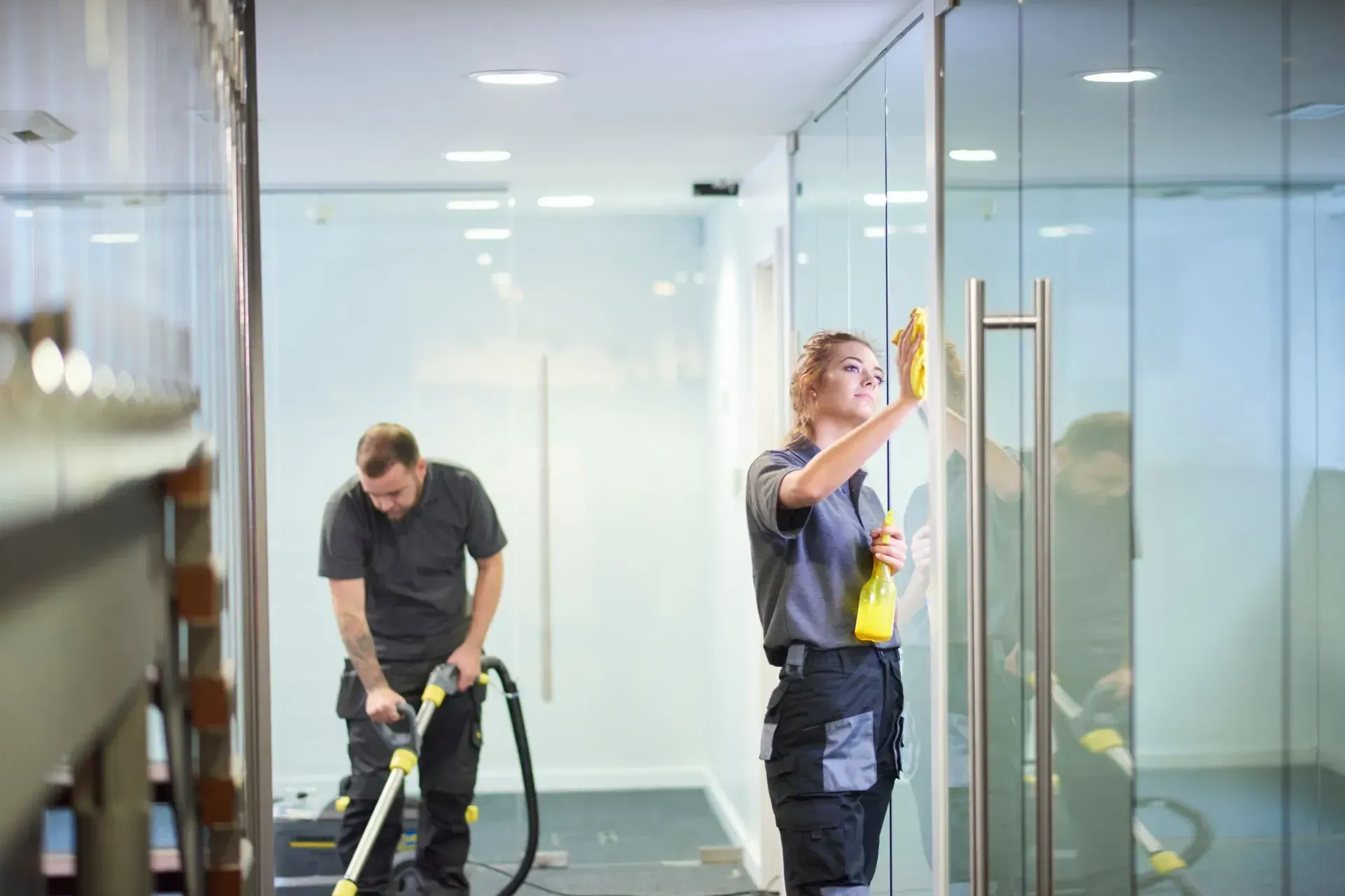 Office Cleaning-A woman cleans a glass door with a cloth and spray bottle, while a man vacuums the floor in an office setting. The scene conveys diligence.