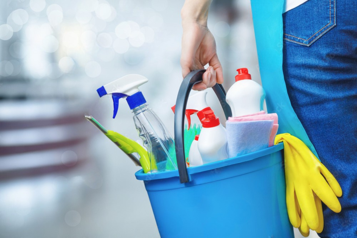 A person holding a blue bucket filled with cleaning supplies, including spray bottles and gloves.