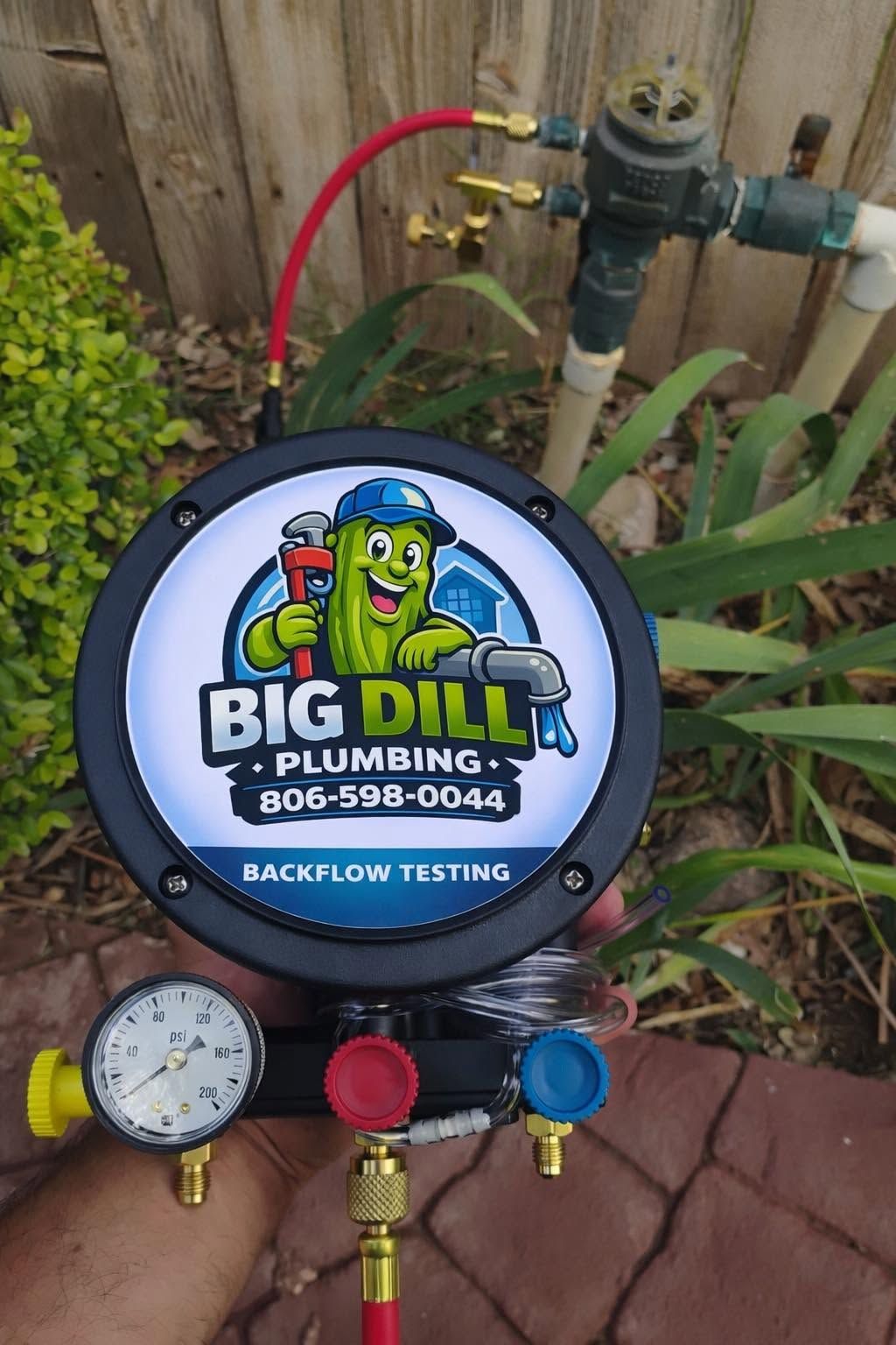 Big Dilli Plumbing backflow testing unit with gauges, red and blue valves, beside a garden fence.