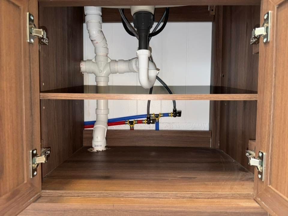 Open cabinet under sink with white plumbing pipes and wooden shelves inside.
