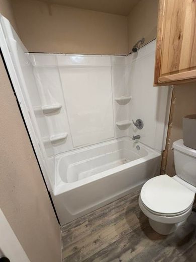 Small bathroom with a white bathtub/shower combo, toilet, wood-pattern floor, and wall cabinet.