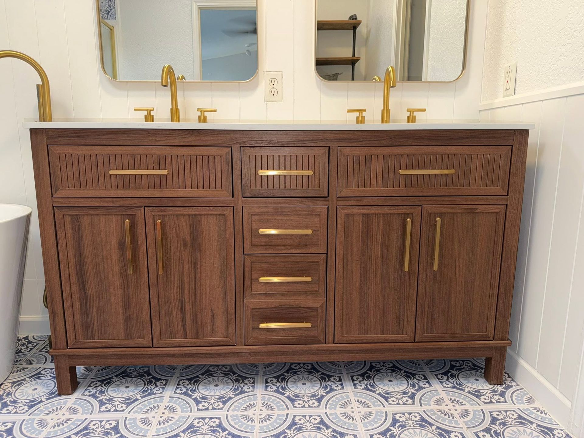 Walnut double vanity with gold faucets and mirrors in a white bathroom with patterned tile floor