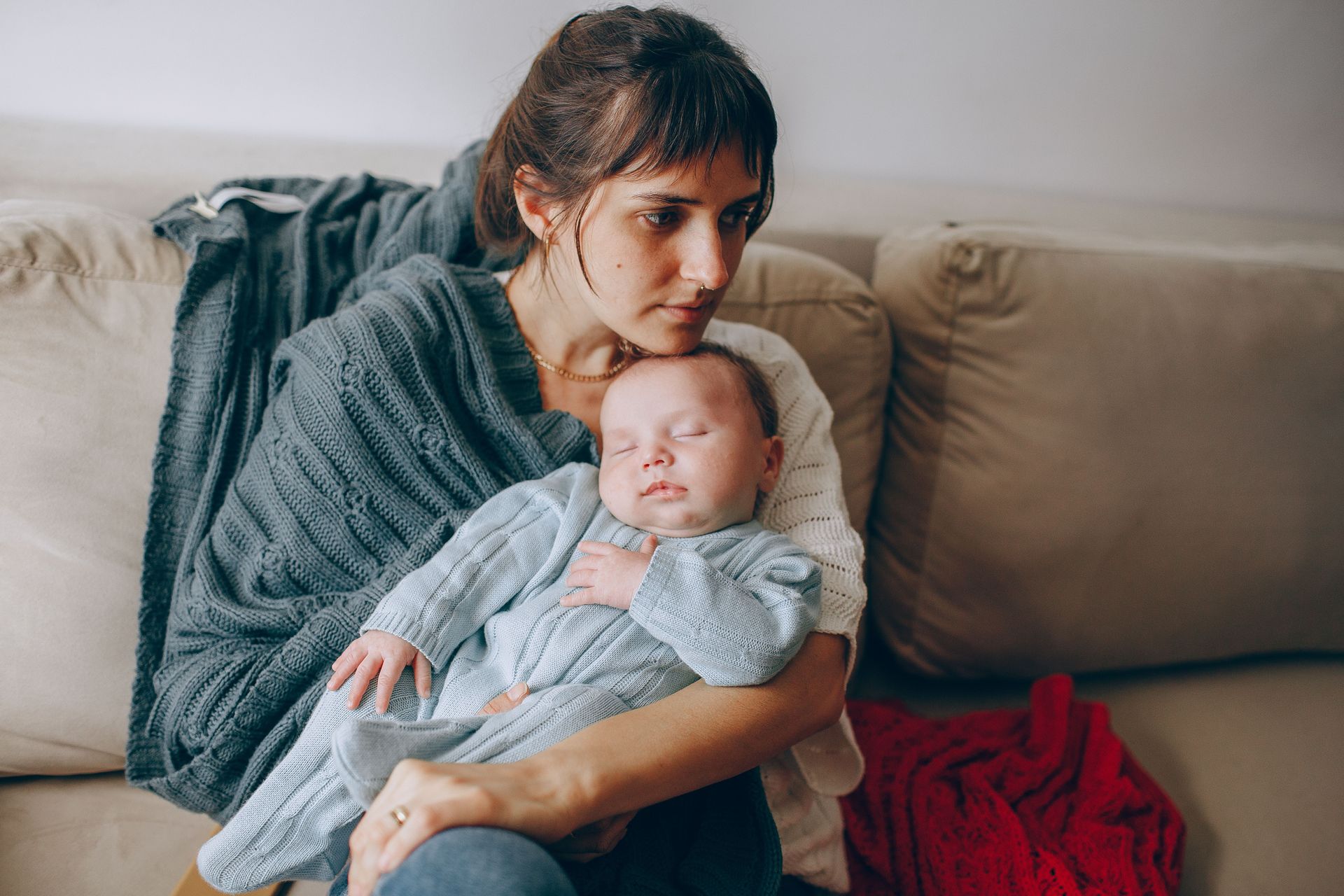 Postpartum woman with depression or anxiety holding a sleeping baby on a beige couch, with a red blanket beside them.