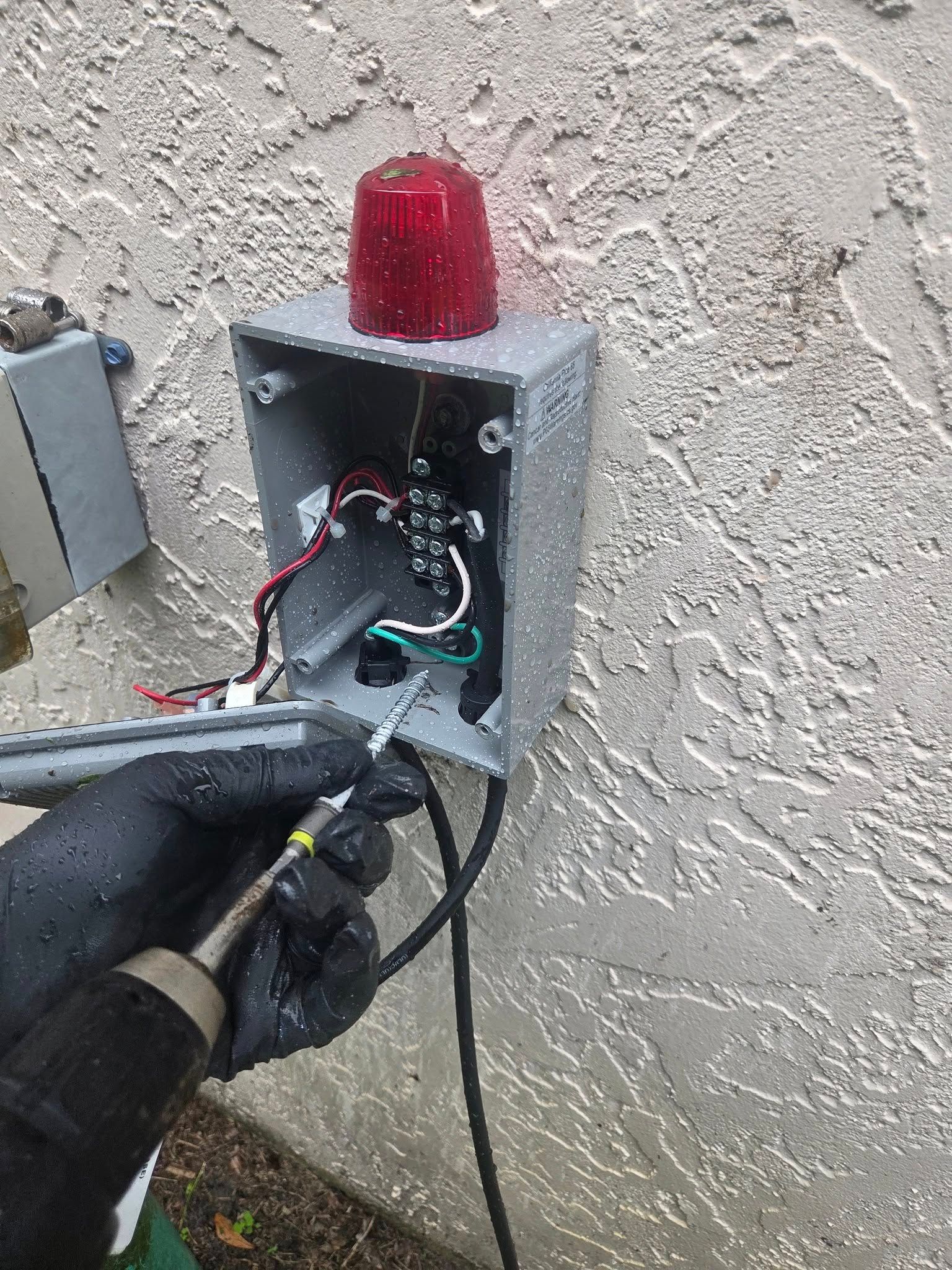 Person in black gloves using a drill on electrical box on stucco wall. Red light on top.