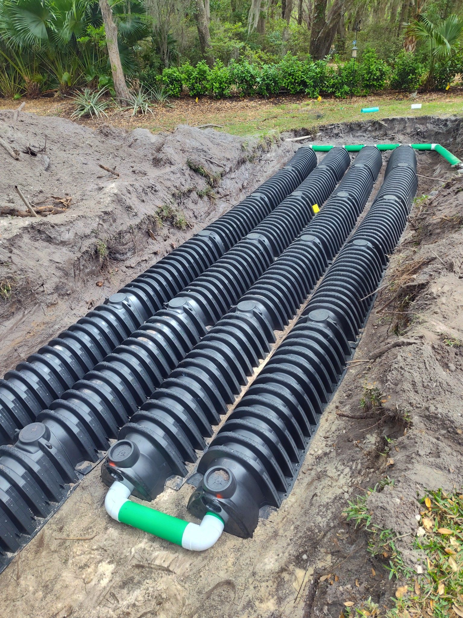 Black plastic septic system components in a trench, connected by green and white pipes, outdoors.