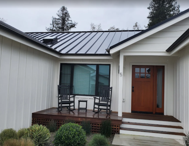 White house with dark metal roof, wood door, porch, two rocking chairs, and greenery.