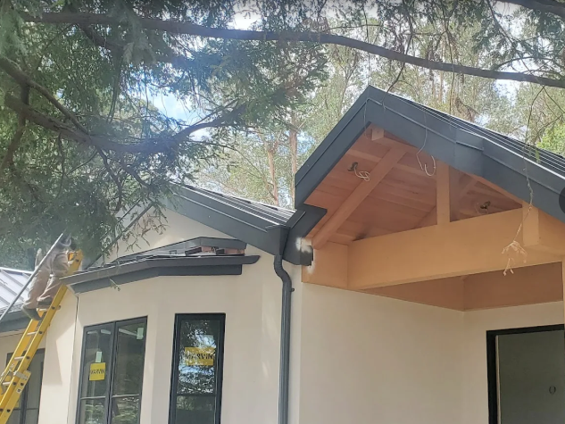 Exterior view of a house under construction with a dark roof and trim, and exposed wooden beams.