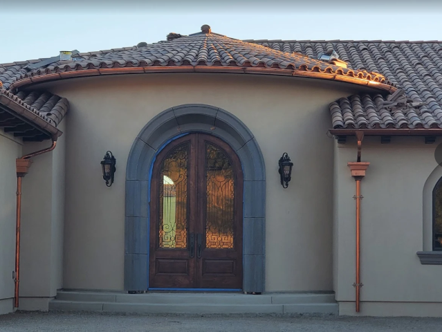 Tan stucco house entrance with arched double doors, dark stone trim, copper gutters, and sconces.