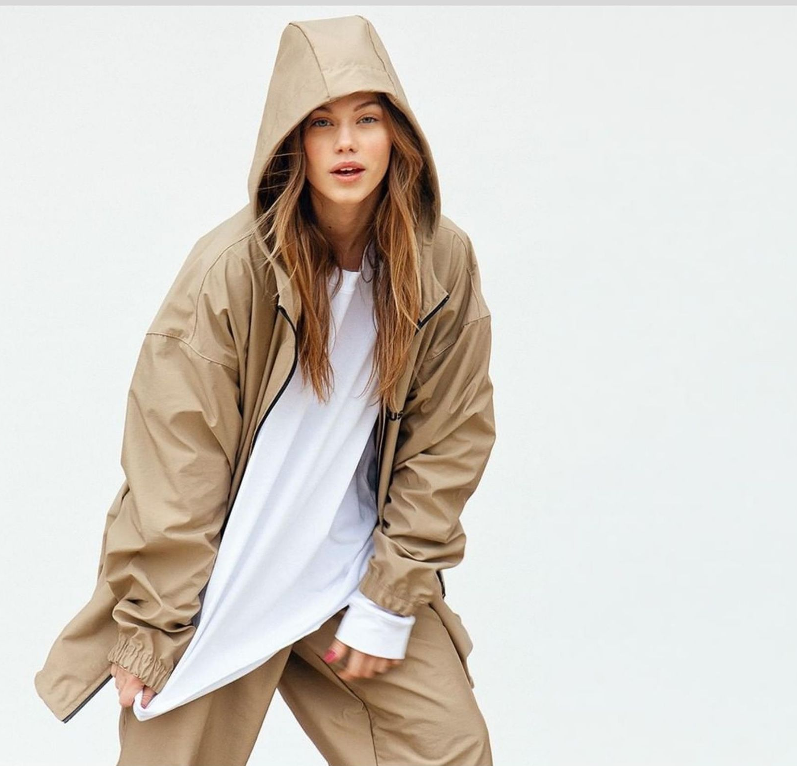 Persona che indossa una giacca con cappuccio color marrone chiaro sopra una camicia bianca e pantaloni color marrone chiaro, in posa su uno sfondo bianco.