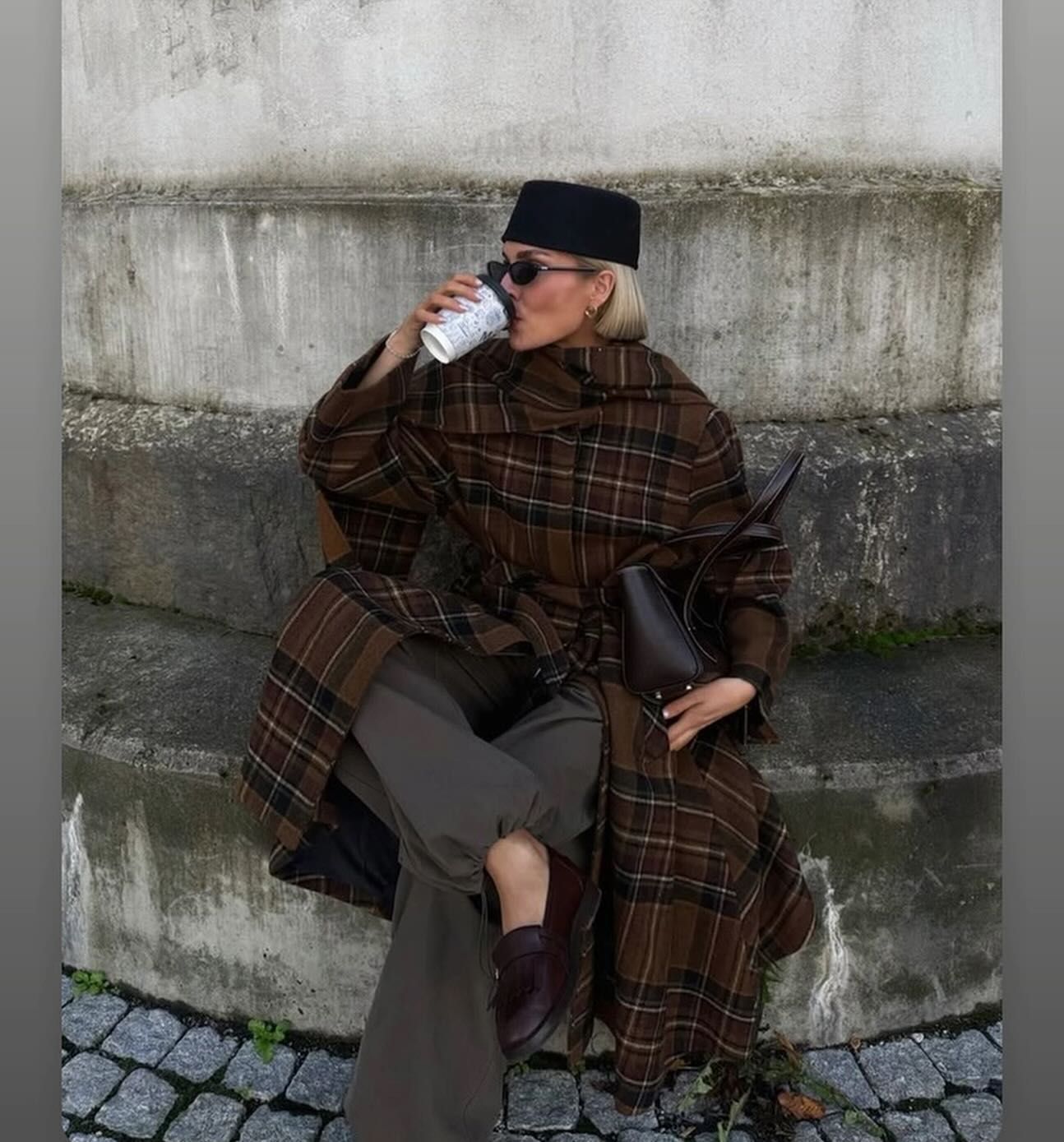Persona seduta all'aperto che sorseggia un caffè, indossa un cappotto scozzese marrone, pantaloni scuri e un cappello nero.