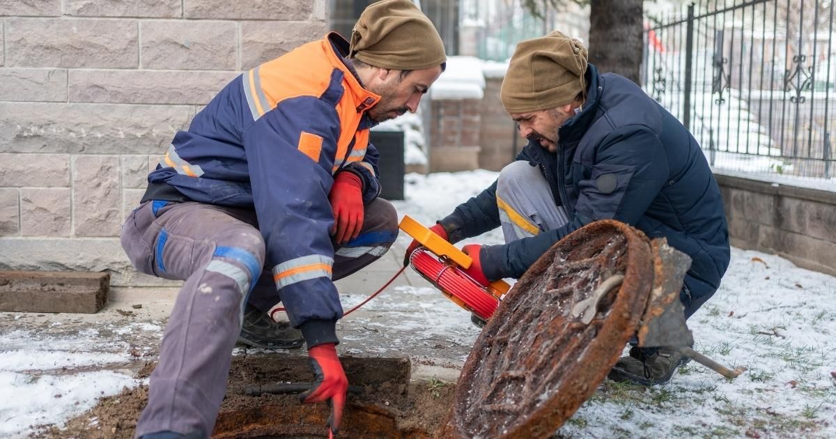 Two utility workers examine a manhole on a snowy street. One kneels, the other holds equipment.