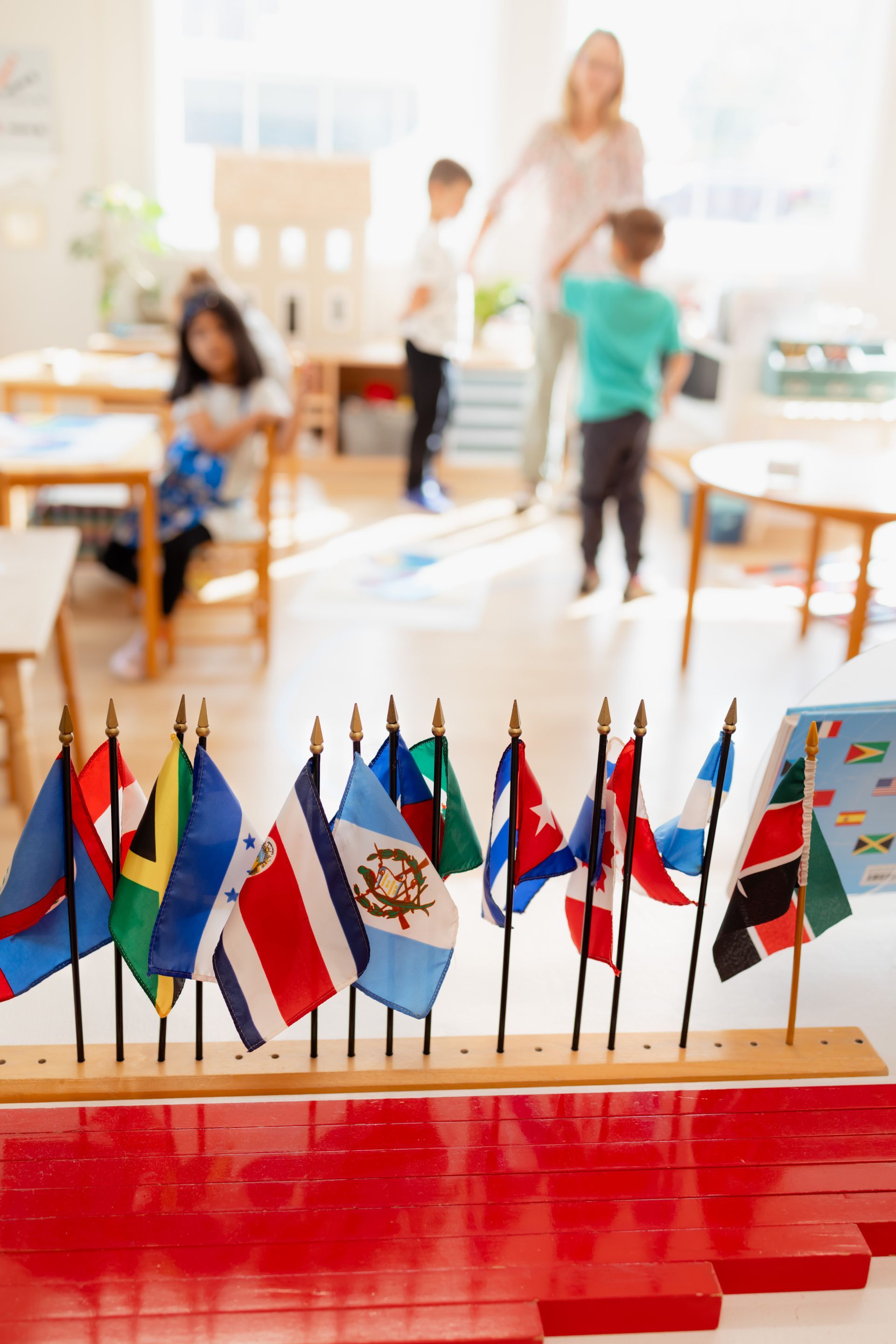Row of colorful miniature flags on a classroom table with children and a teacher blurred in the background