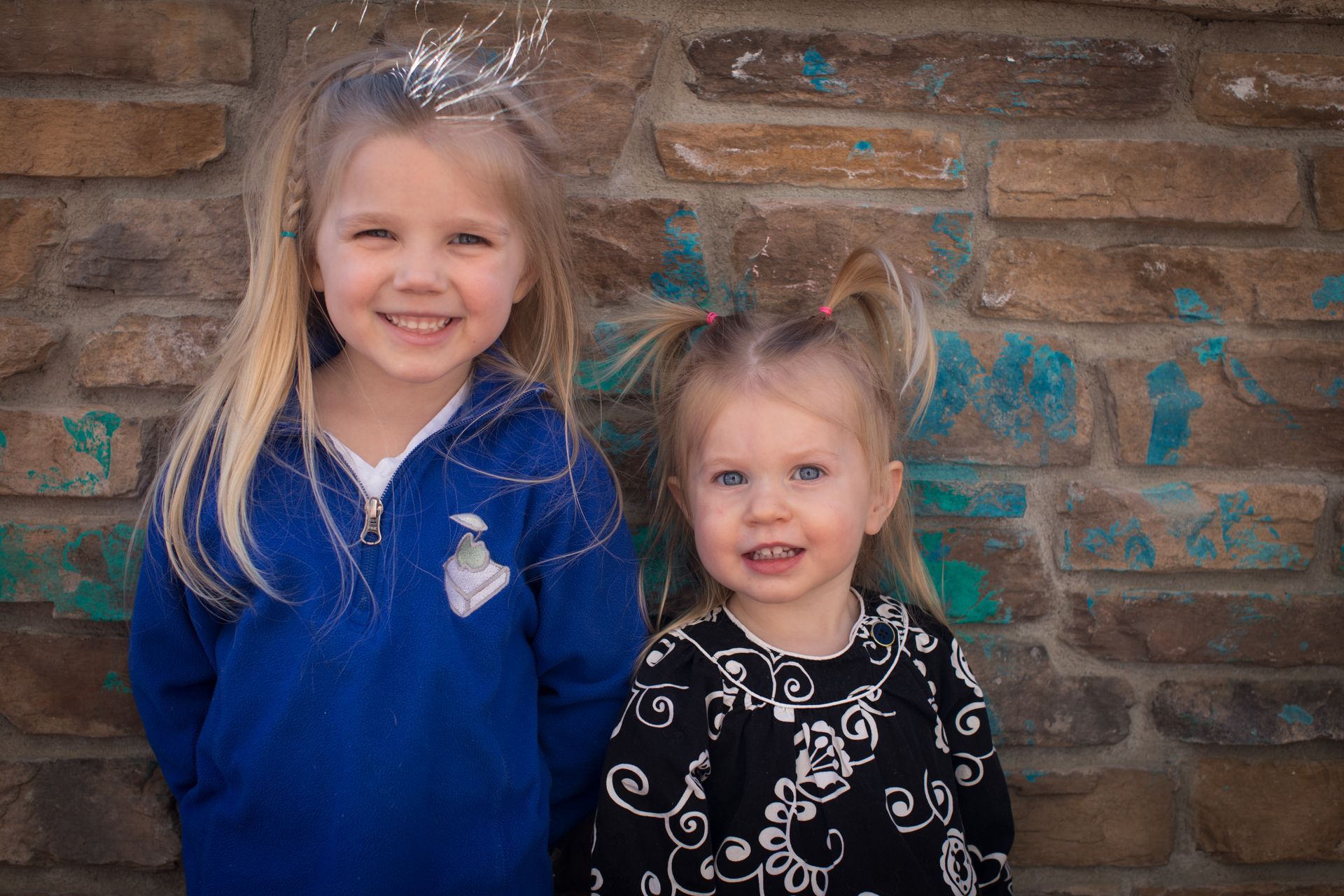 Two smiling children in blue and black outfits standing against a brick wall