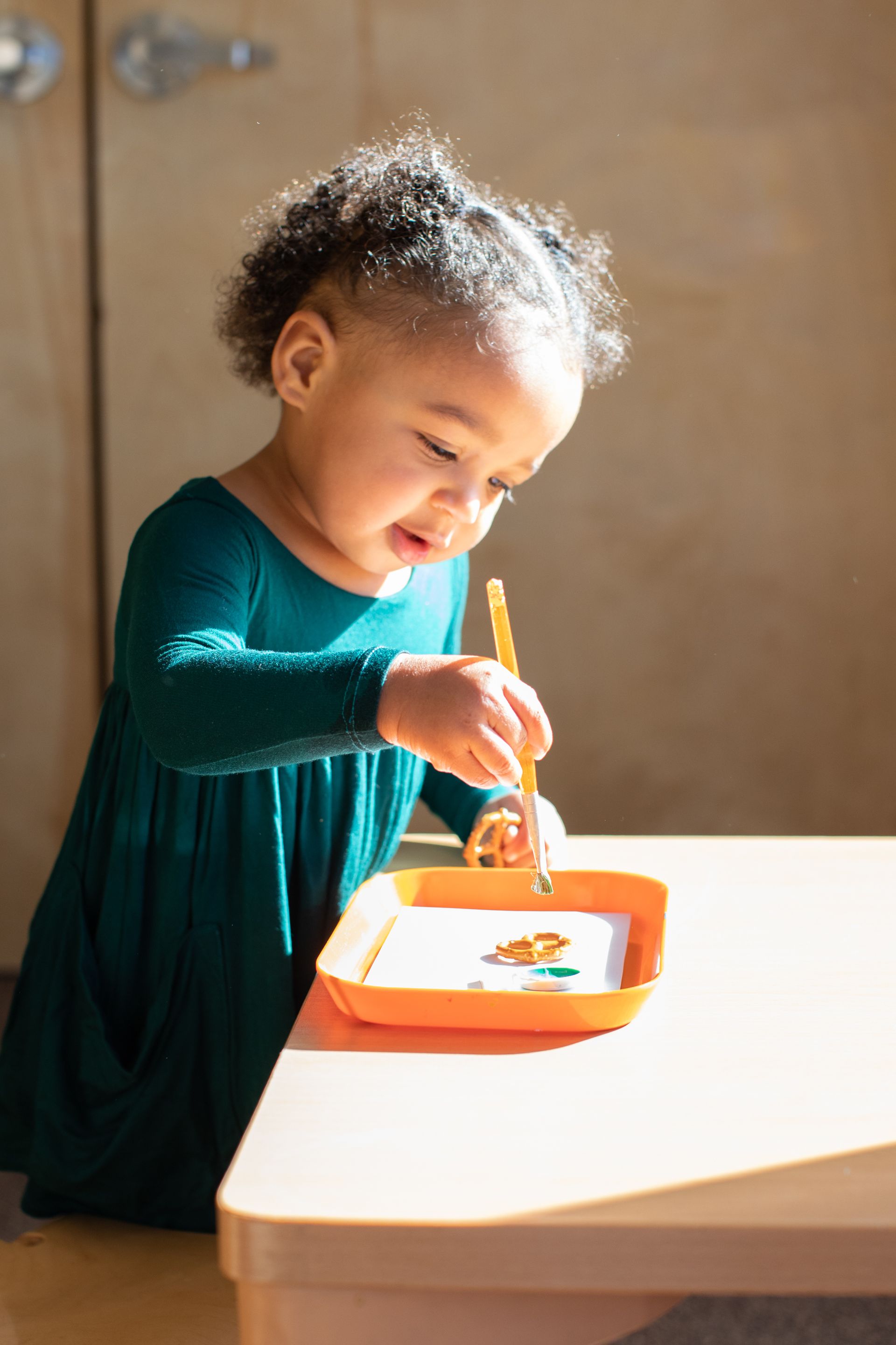 Toddler in a teal dress scooping food from an orange tray at a table.