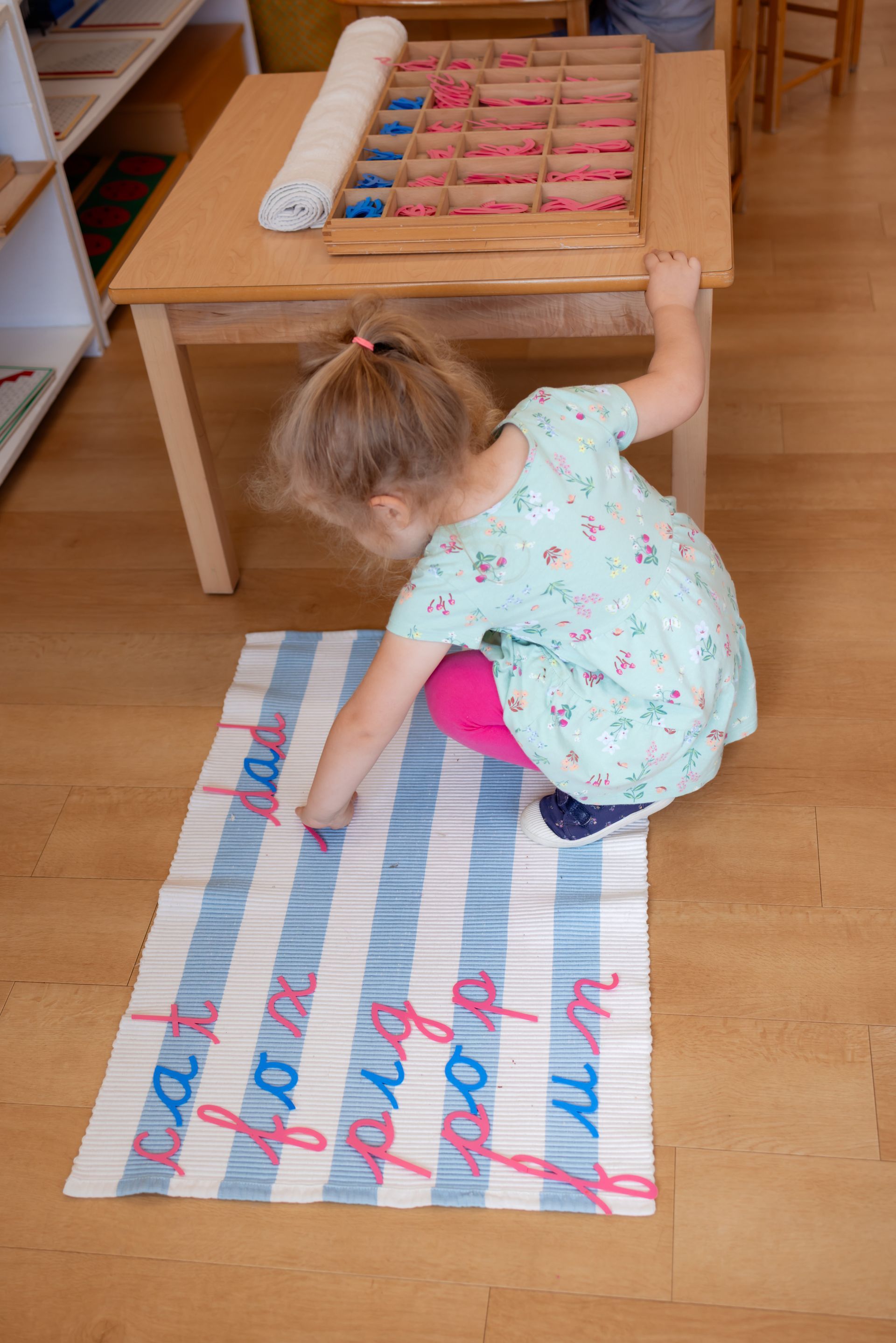 Child kneeling on a striped rug, reaching toward a low wooden shelf in a playroom.