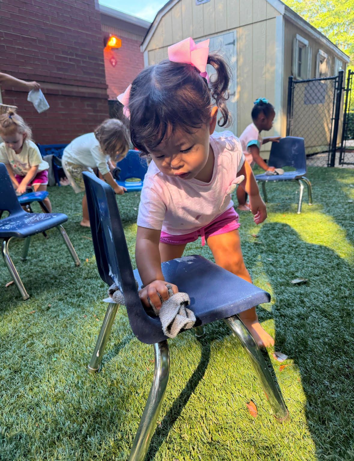 Child washing a small chair outdoors on grass, wearing a pink bow and white shirt.