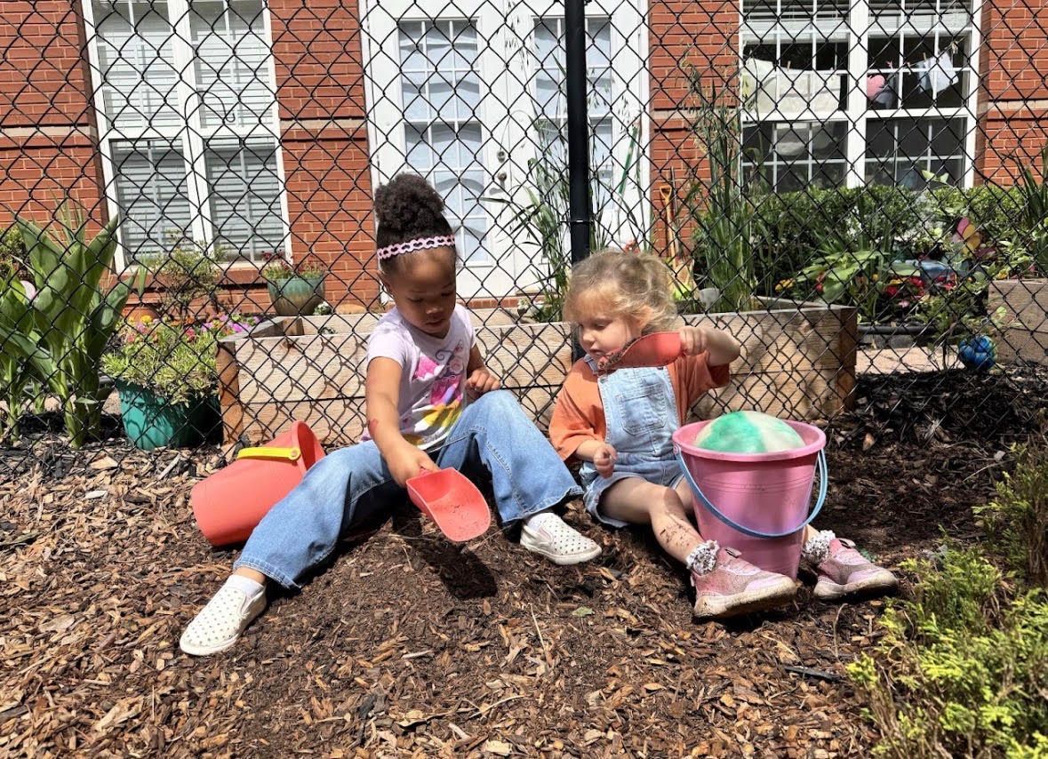 Two children sitting on mulch near a brick building, playing with buckets and garden tools.