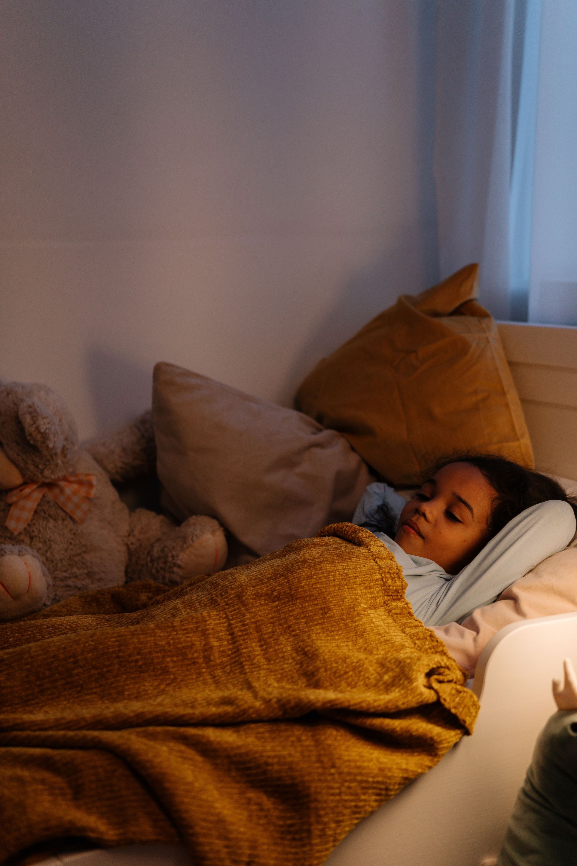 Person sleeping under a mustard blanket on a couch with pillows in a softly lit room