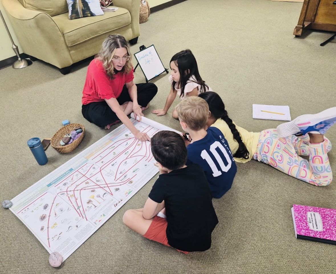 Child doing work at a table while a teacher assists, with another child working on floor.