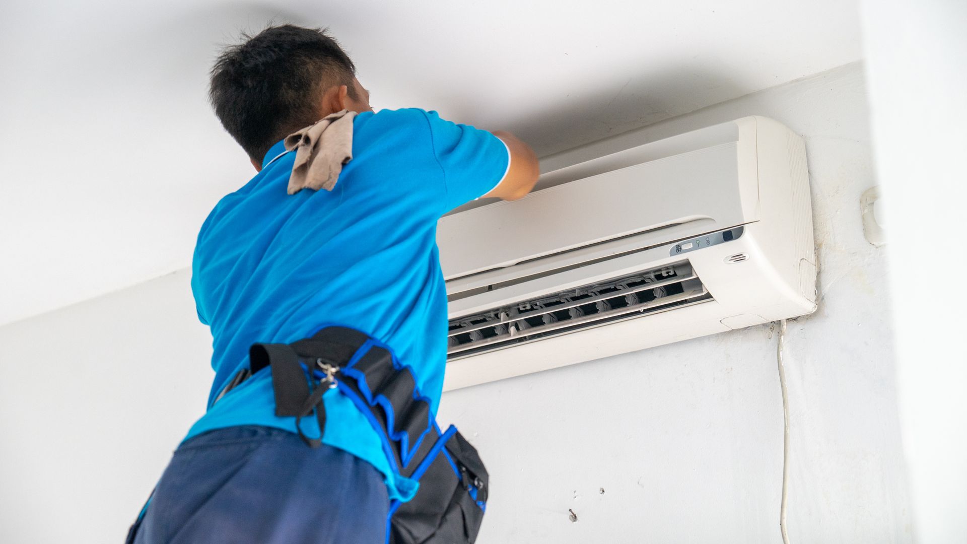 Technician in blue uniform inspecting a wall-mounted air conditioner while on the phone