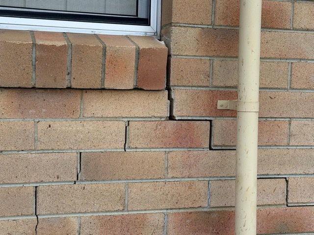 Cracked brick wall beneath a window with a vertical pipe in front