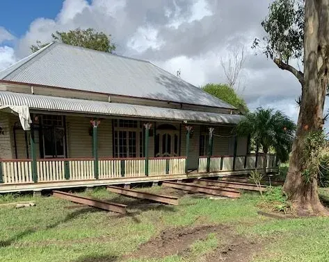 Old wooden house with a porch and tin roof in a grassy yard, with a large tree in front.