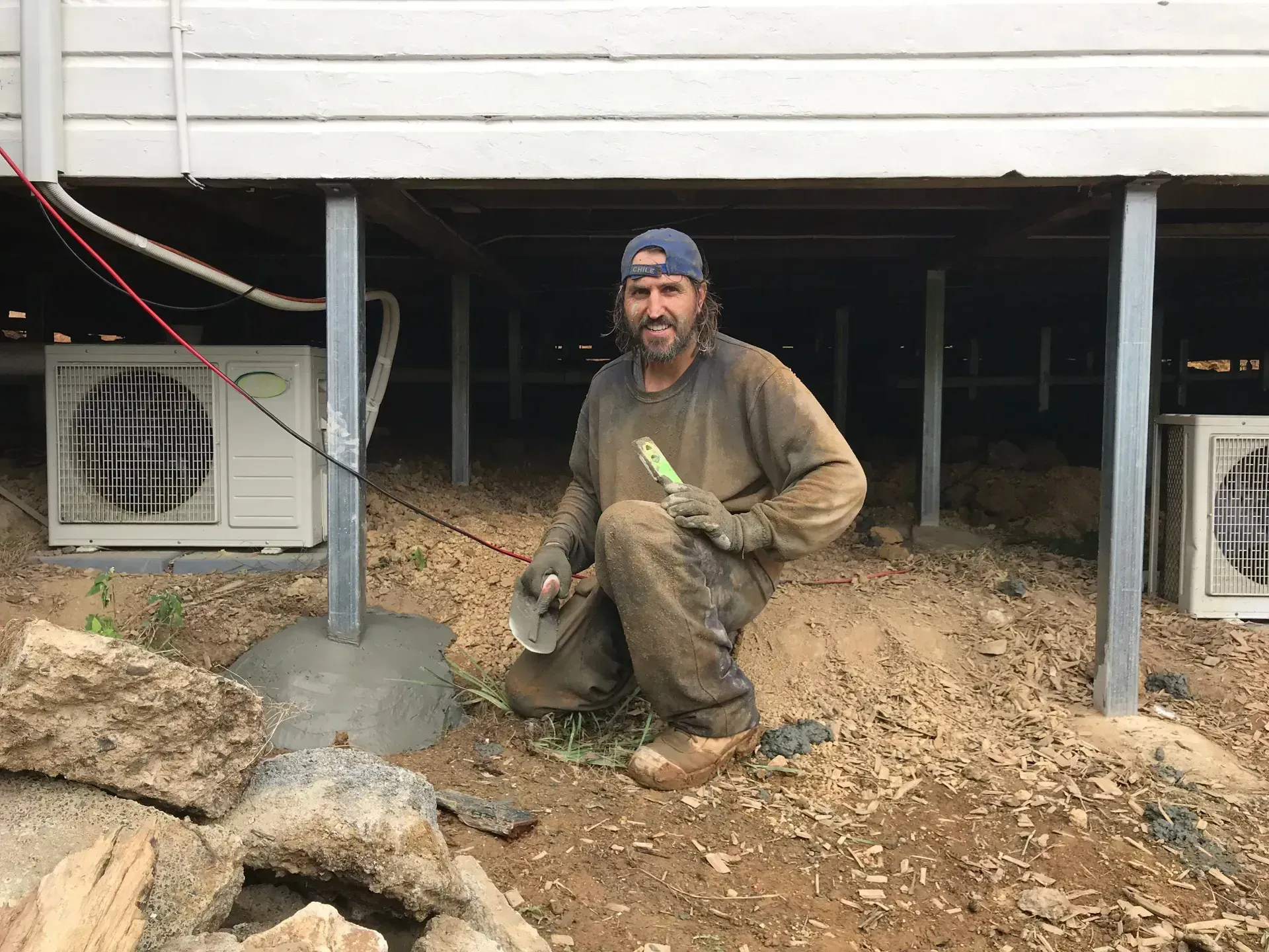 Muddy worker crouches under a raised house beside debris and support posts, with window AC units visible.