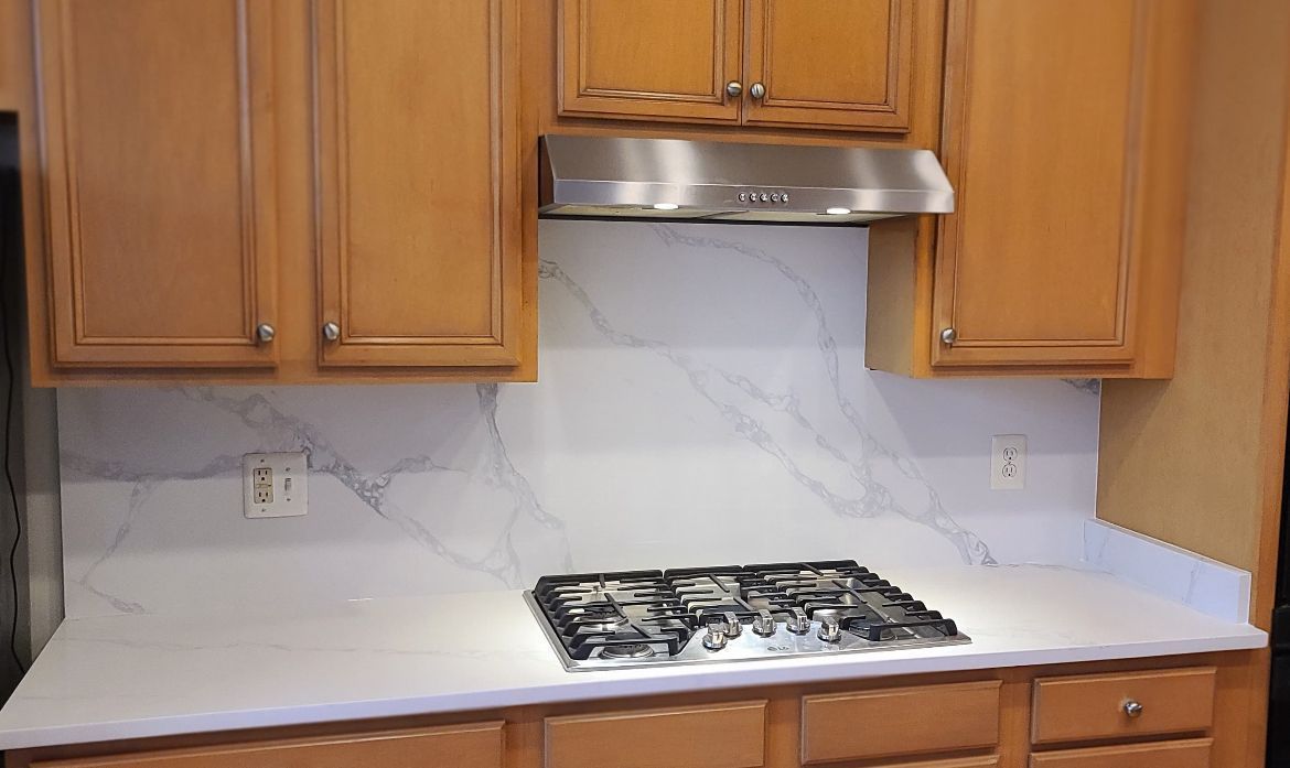 Kitchen with light wood cabinets, white marble backsplash and countertop, stainless steel range hood and gas stove.