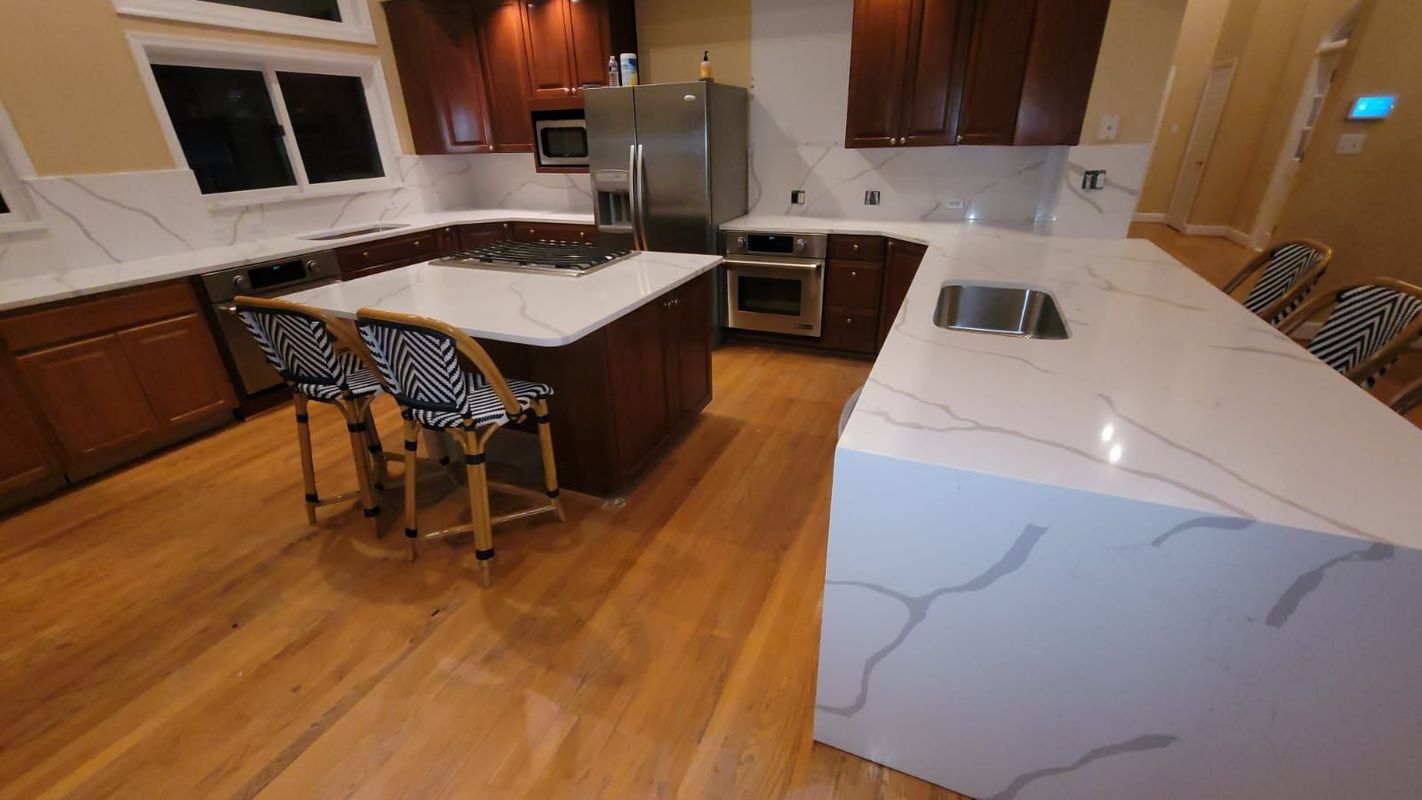 A modern kitchen with wood floors, white countertops, and dark brown cabinets. Two black and white patterned bar stools sit at the island.