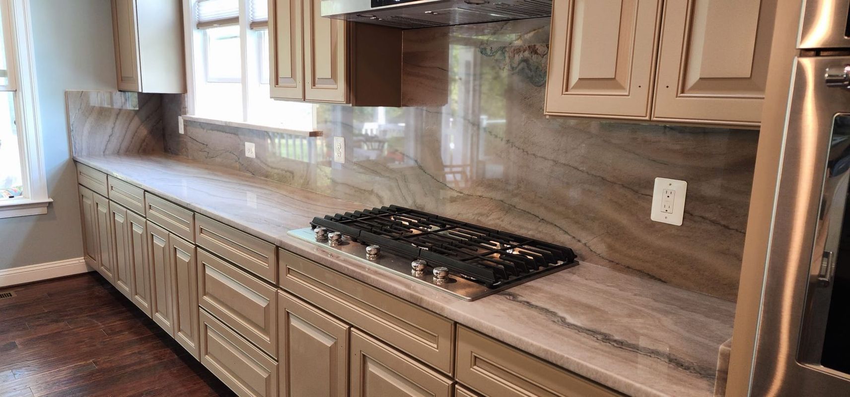 Kitchen with light-colored cabinets, marble countertops, stovetop, and window.