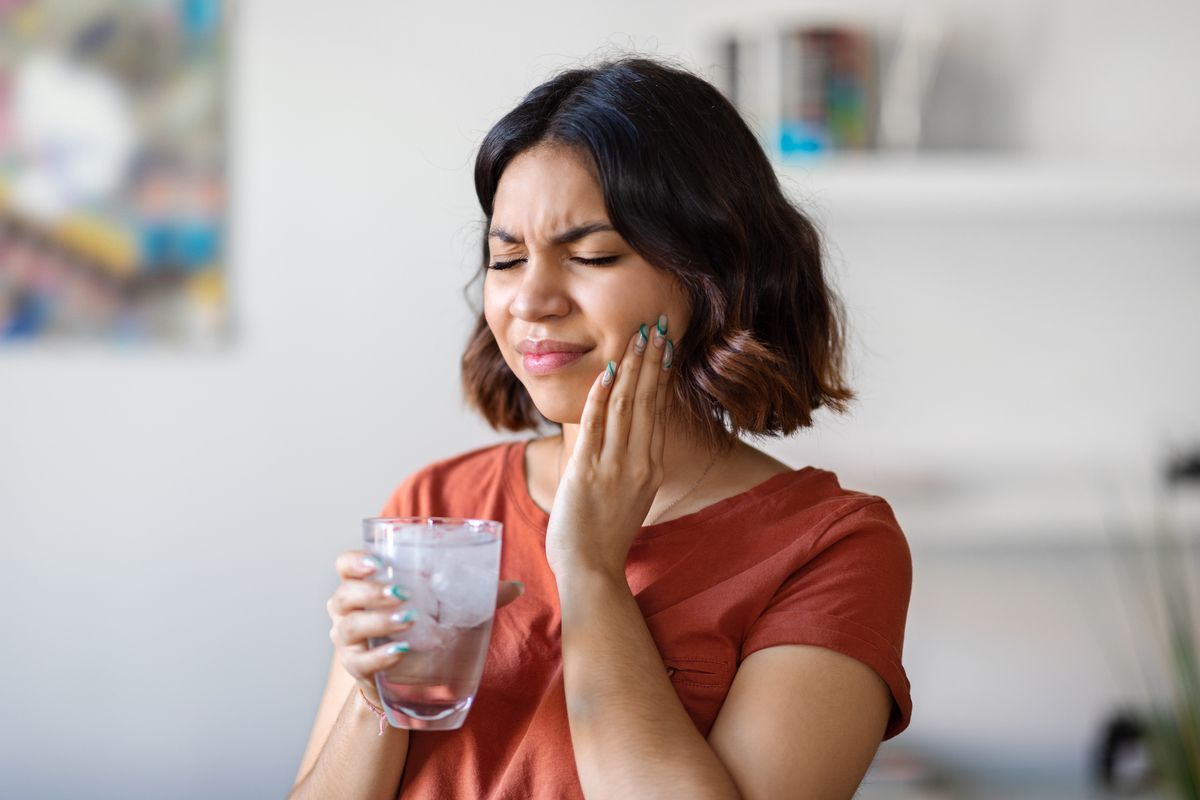 Woman holding a glass with ice, grimacing, touching her cheek in discomfort, appearing to have a toothache.