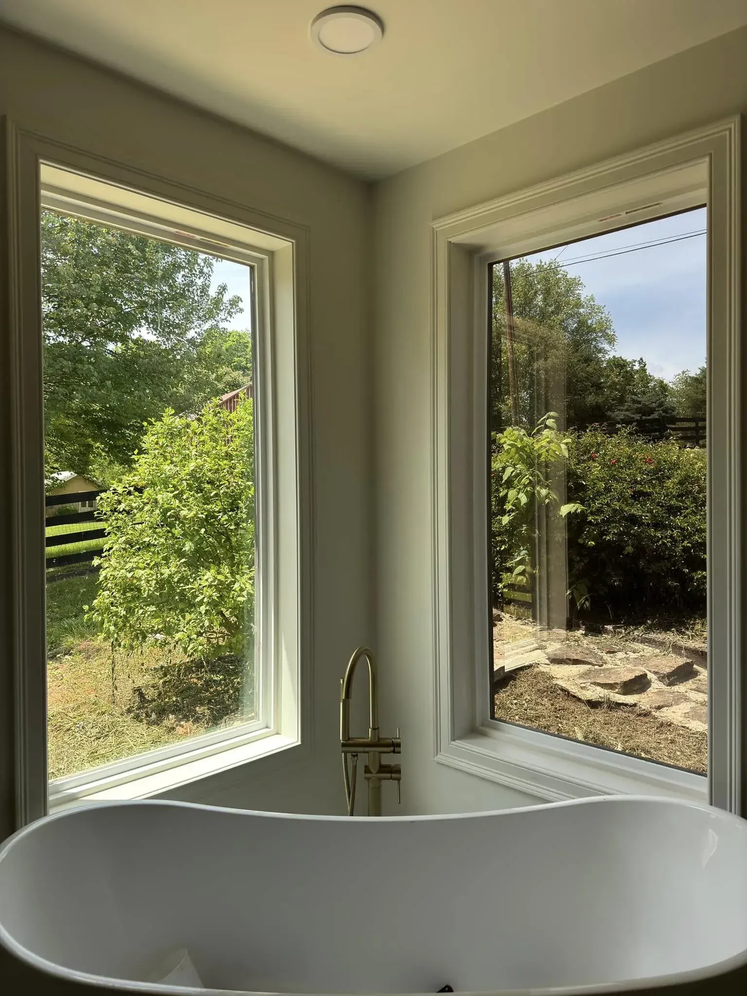 Bathroom with a white tub between two windows overlooking a lush green yard.