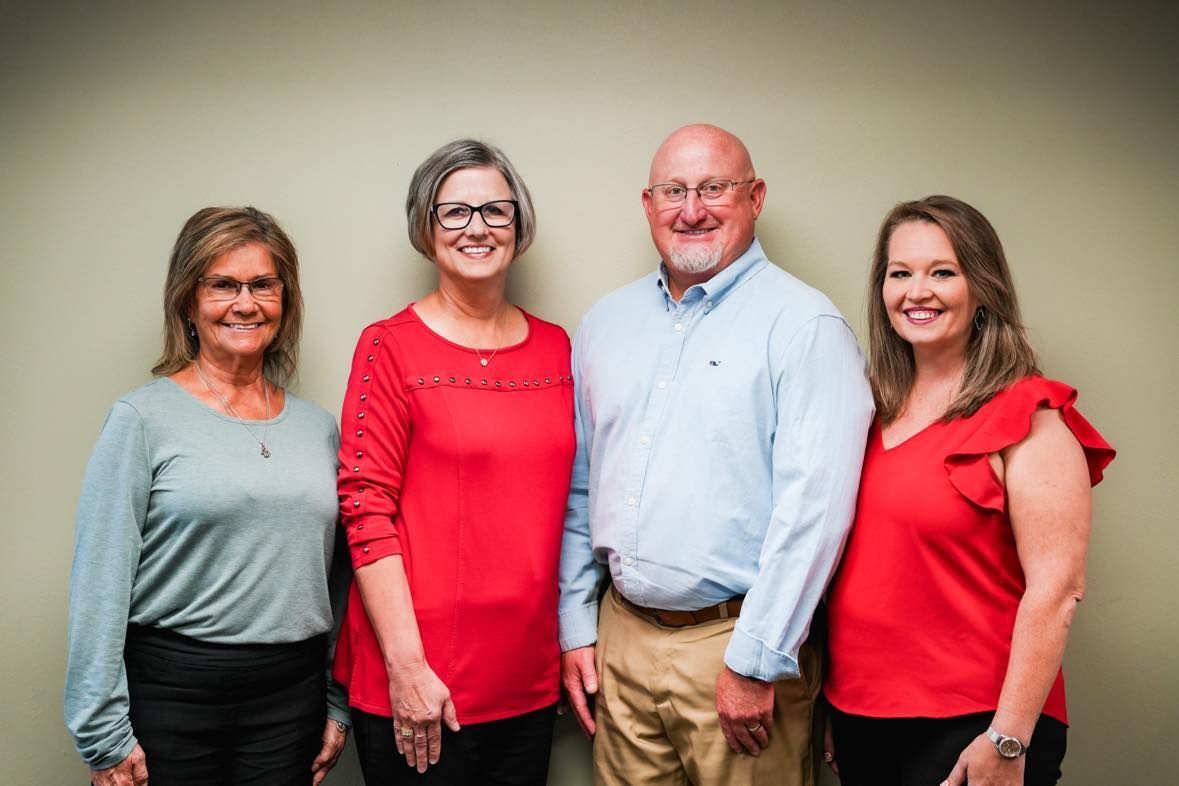 Four people stand smiling against a gray wall, wearing red, blue, and gray tops.