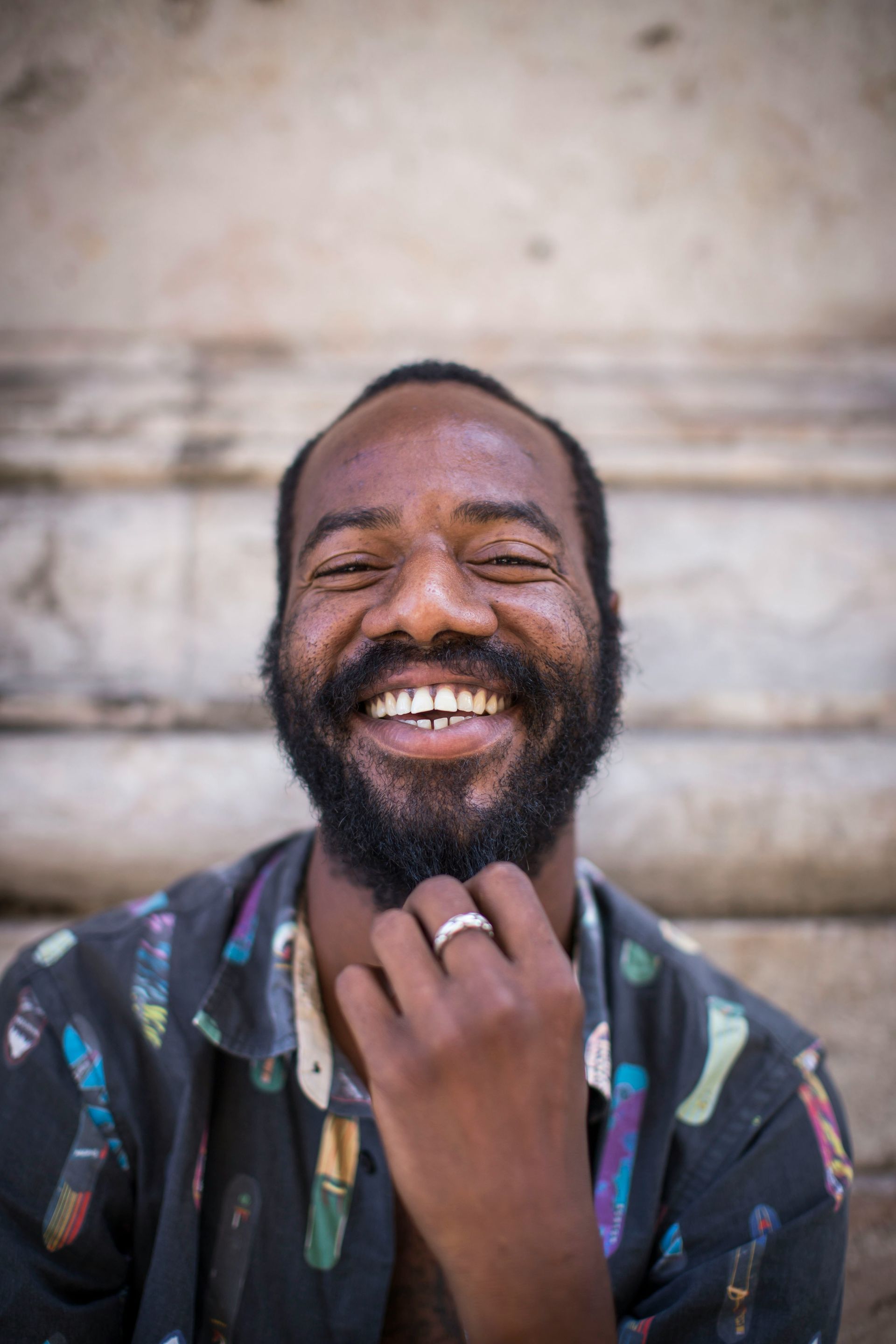 A Black man with a full beard smiles broadly, hand to his chin, in front of a stone structure.