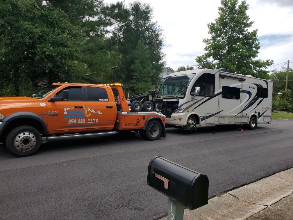 Orange tow truck pulling a damaged white RV on a roadside under cloudy skies.