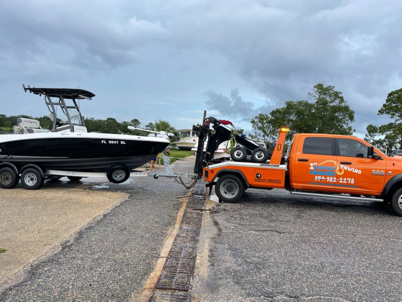 Orange tow truck pulling a black boat on a trailer along a gravel road under cloudy skies