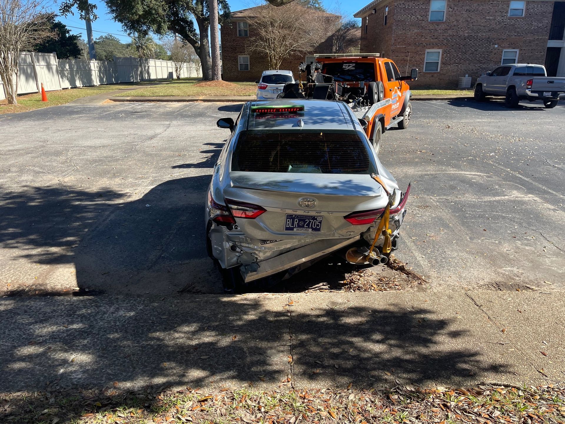 Damaged silver sedan with severe rear-end crash damage on a gravel lot, with an orange tow truck behind it