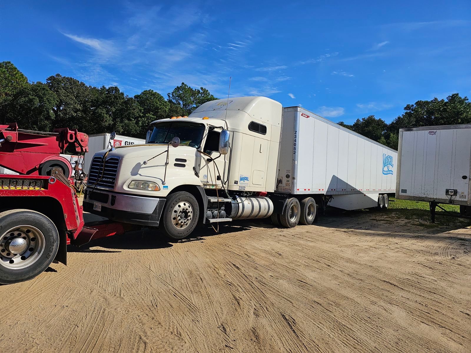 White semi-truck with trailer parked on dirt lot beside a red truck under a blue sky
