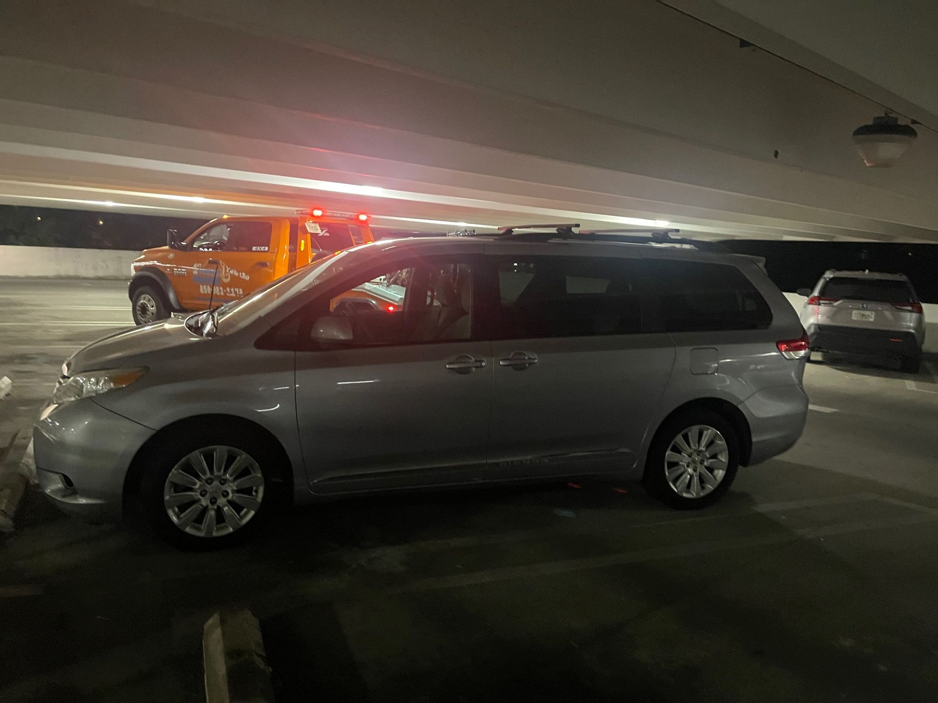 Silver minivan parked in a dim garage beside an orange tow truck with flashing lights