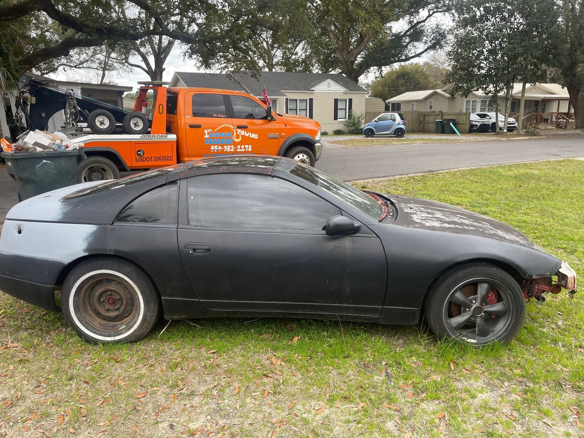Black sports coupe parked on grass with orange tow truck behind it in a suburban neighborhood